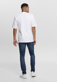 White cotton t-shirt with a relaxed fit, paired with dark blue denim jeans. Model stands casually, facing slightly away from the camera.