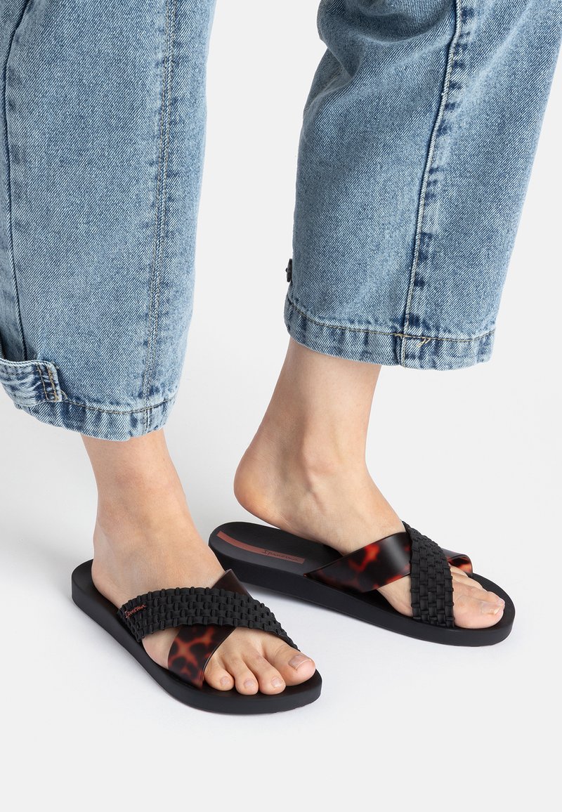 Schwarze Sandalen mit Kreuzrippen und einem geflochtenen Design, verziert mit Schildpatt-Akzenten. Sie besitzen eine flache Sohle und eine offene Zehenpartie und liegen auf hellem blauen Denim.