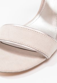 Gros plan d'une bride de sandale en daim beige avec un détail de couture sur la zone des orteils et une semelle intérieure métallique.