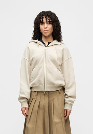 Beige lynlåstrøje i blødt stof med snøre ved hætten, løs pasform og forlommer, kombineret med et plisseret brunt skørt.