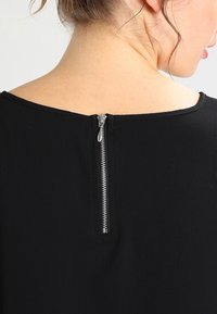 Svart topp med en enkel rund halsringning. En silverfärgad dragkedja löper ner längs den centrala bakdelen, vilket kontrasterar med den släta texturen på tyget.