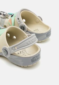 Crocs in light beige with mint accents, featuring a gray strap embossed with "Star Wars." Interior displays character graphic. Lightweight design.