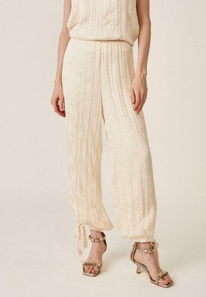 Mujer vistiendo pantalones arrugados color crema con lazos en los tobillos y sandalias de tacón alto beige adornadas con decoraciones florales.