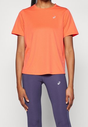 Woman wearing orange short-sleeve sports shirt and purple leggings, both with white ASICS logos, standing against a plain background.