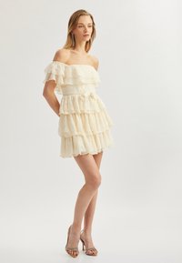 Kreemi off-the-shoulder kleit kihiliste volangide ja pitsitükiga, millel on vormitud vöökoht ja paelteade. Kantud rihmadega kontsakingadega.