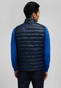 Marineblaue Steppweste mit horizontalem Steppmuster, hohem Kragen und dezentem Logodetail. Über einem blauen Langarmshirt getragen.