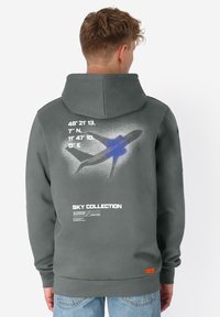 Jonge man in een grijze hoodie met een blauwe vliegtuigafbeelding en witte coördinaten op de rug, gecombineerd met lichtblauwe jeans.