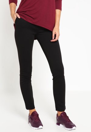 Leggings noirs au design ajusté, texture lisse et taille élastique, associés à des chaussures de sport violettes foncées avec des accents blancs.