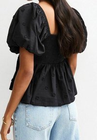 Blouse en coton noir à manches bouffantes, ornée de broderies florales. Le dos présente un design smocké, associé à un jean taille haute bleu clair.