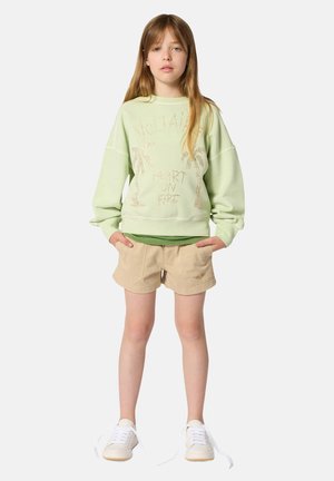 Jeune fille debout, les mains dans les poches, portant un sweat-shirt vert clair avec un motif de palmier, un short beige et des baskets blanches aux lacets défaits.
