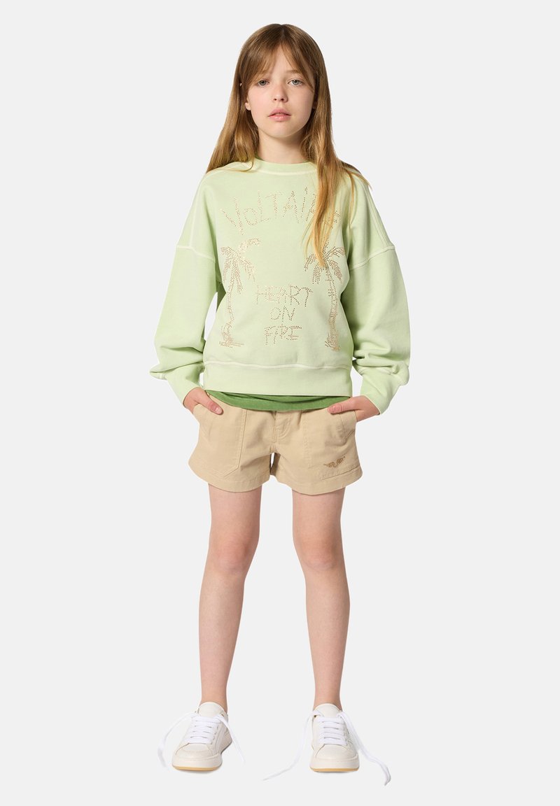 Jeune fille debout, les mains dans les poches, portant un sweat-shirt vert clair avec un motif de palmier, un short beige et des baskets blanches aux lacets défaits.