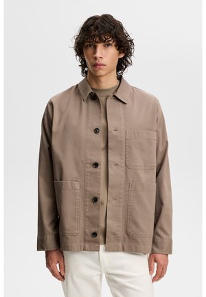 Giovane uomo con capelli ricci e scuri che indossa una giacca taupe con bottoni sopra una camicia abbinata e pantaloni bianchi, in piedi davanti a uno sfondo semplice.