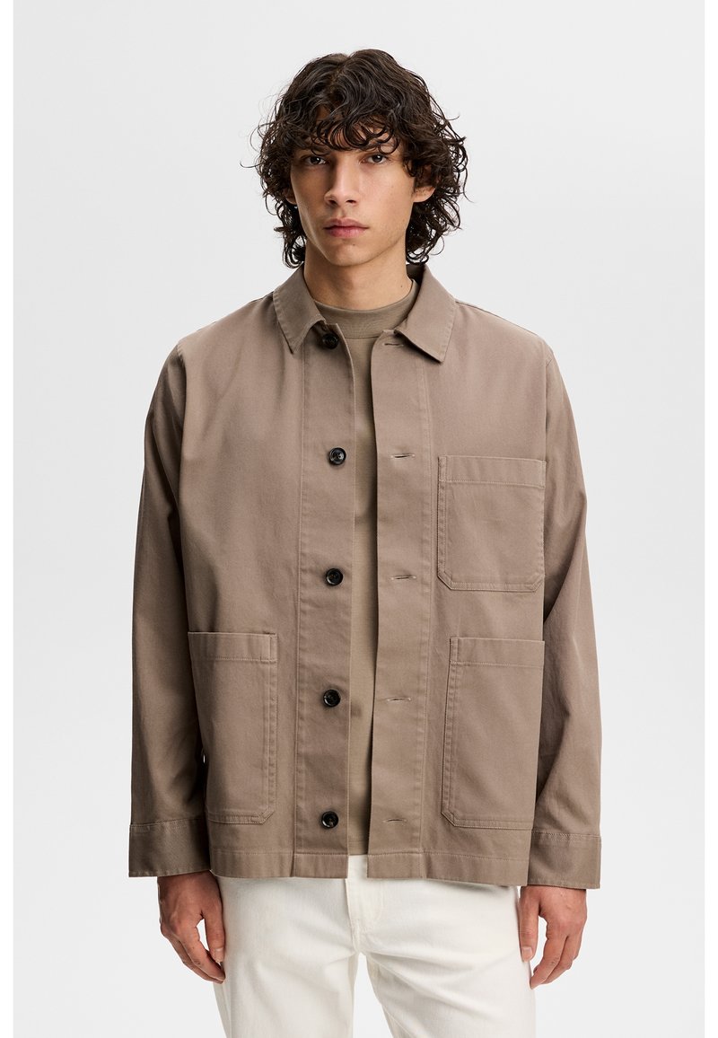 Młody mężczyzna o kręconych ciemnych włosach, ubrany w taupe'ową zapinaną na guziki kurtkę na koszulę w tym samym kolorze i białe spodnie, stojący na jednolitym tle.