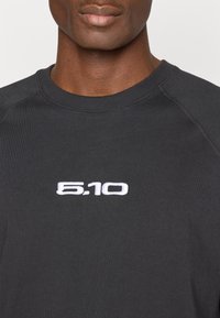 Čierne bavlnené tričko s okrúhlym výstrihom a raglánovými rukávmi, ozdobené bielym vyšívaným logom "5.10" na hrudi.