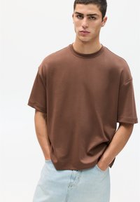 Hnědá oversized tričko s kulatým výstřihem a krátkými rukávy, vyrobené z jemné látky, v kombinaci se světle modrými džíny šortkami. Čisté pozadí.