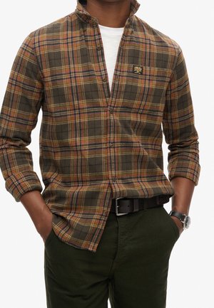 Mann trägt ein braun-orange-schwarzes kariertes Hemd über einem weißen T-Shirt, mit grüner Hose, schwarzem Gürtel und Armbanduhr, Hände in den Taschen.