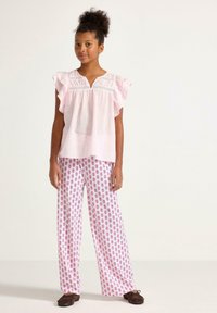 Ljust rosa kortärmad blus med volanger och spetsdetaljer, tillsammans med vita vida byxor med ett rosa trädmönster. Bruna slip-on skor.
