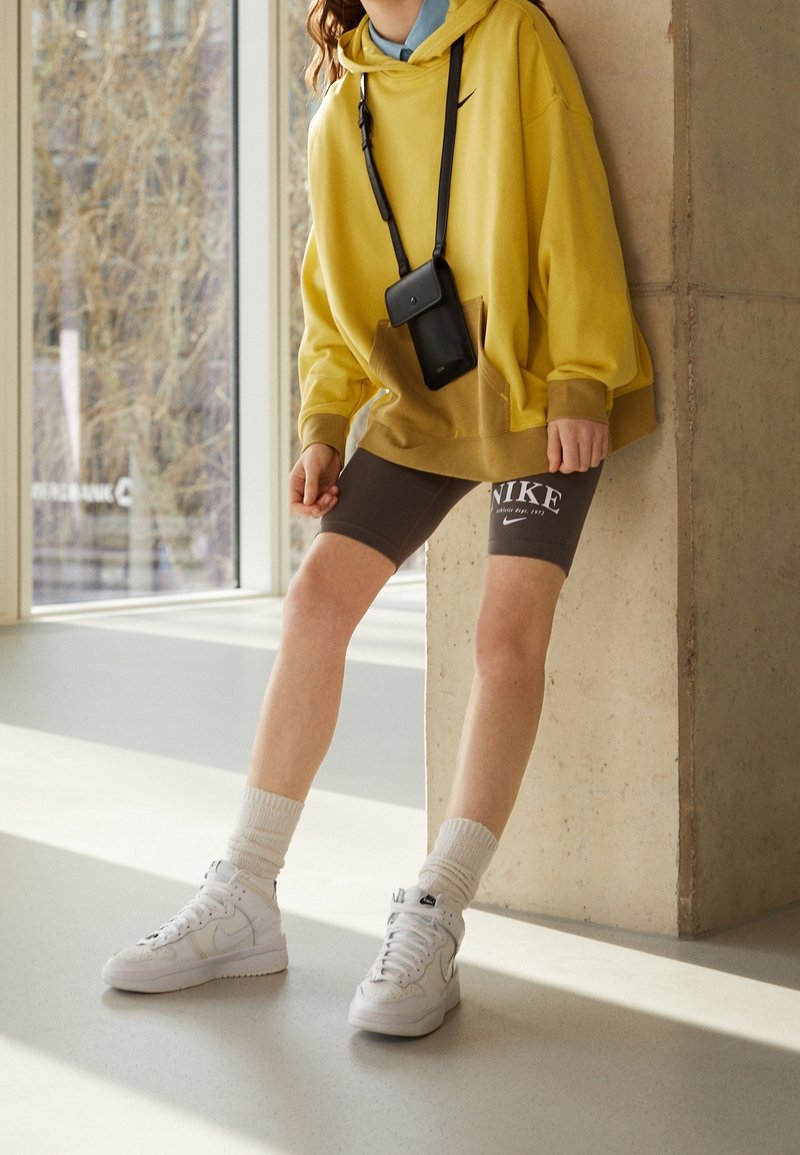 Gul oversized huvtröja, bruna Nike-shorts med logotyp, vita högklackade sneakers och ljusa strumpor. Enkel svart crossbody-väska. Minimalistisk bakgrund.