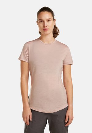 Ruskeatukkainen nainen poninhännällä, pukeutunut vaaleanpunaiseen lyhythihaiseen paitaan ja tummanharmaisiin housuihin, seisoo yksinkertaista valkoista taustaa vasten.