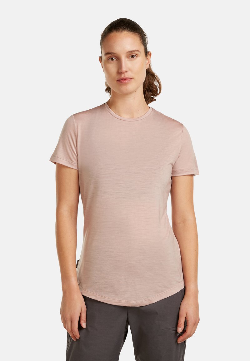 Woman with brown hair in a ponytail wearing a light pink short-sleeve shirt and dark gray pants, standing against a plain white background.