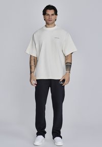 Lielizmēra krēma kokvilnas t-krekls ar īsām piedurknēm un apaļu kakla izgriezumu, savienots ar melniem sporta biksēm ar izšūtu logotipu un baltām sporta apaviem.