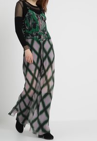 Femme portant un haut noir transparent à manches longues sous une robe maxi sans manches verte et rose à motif avec une fermeture éclair devant, assortie à des bottines noires.