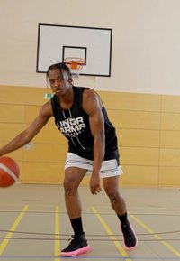 Basketball player in a black sleeveless shirt with white accents, black shorts, and black/pink sneakers, dribbling a basketball indoors.
