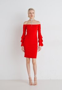 Rød avskjæringskjole med flagrende ermer, tettsittende silhuett, glatt tekstur og knelange design. Båret med hvite sandaler med remmer.