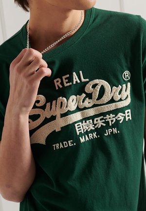 Person iført mørkegrøn skjorte med hvidt tekstureret "Real SuperDry"-logo og japansk tekst, der holder en sølvkæde nær kraven.