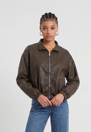 Jeune femme aux cheveux tressés portant des boucles d’oreilles créoles dorées, une veste zippée marron foncé et un jean bleu, debout devant un fond uni.