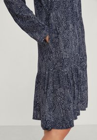 Abito navy con un motivo testurizzato caratterizzato da disegni astratti bianchi. Maniche lunghe, orlo con volant e tasche laterali visibili.