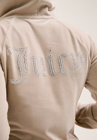 Bež majica s kapuljačom meke teksture, s ukrasnim kristalima koji ispisuju riječ "Juicy" na leđima u kurzivnom fontu.