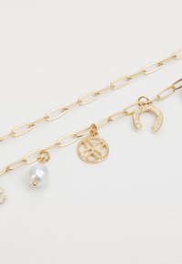 Bracelet en chaîne dorée avec des maillons ovales, un charme en perle et trois pendants décoratifs : une fleur sculptée, un médaillon rond et un fer à cheval.