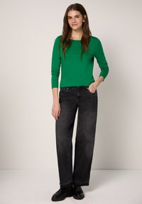 Vihreä pitkähihainen paita yhdistettynä mustiin leveälahkeisiin farkkuihin ja mustiin saappaisiin. Paidassa on pyöreä pääntie, ja farkuissa on etutaskut.