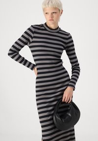 Vestido de manga longa riscado em tecido canelado cinza e preto, com corte justo. Modelo segurando uma bolsa preta em forma de meia-lua.