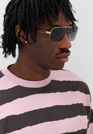 Uomo con dreadlocks corti che indossa occhiali da sole scuri, un piccolo orecchino e una camicia a righe rosa e nere, rivolto verso destra.