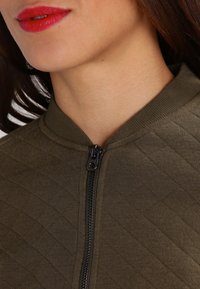 Olivgrön vadderad jacka med hög krage, featuring en framsida med dragkedja och textil med struktur som framhäver en välstrukturerad design.