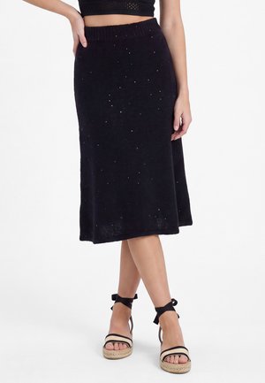 La donna indossa una gonna nera al ginocchio con un motivo leggermente scintillante e sandali con zeppa neri con lacci alla caviglia e cinturini beige.