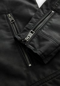 Chaqueta de cuero negra con textura suave, costuras visibles y bolsillos con cremallera en las mangas, con cremalleras y tiradores metálicos.