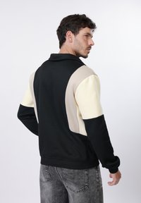 Homme portant un sweat-shirt à manches longues color block noir, beige et crème, tourné de dos, avec un jean gris délavé sur fond blanc.