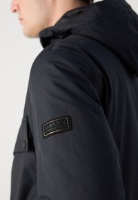 Veste bleu marine avec capuche, présentant un tissu texturé. Un patch de logo noir est visible sur le haut du bras, ajoutant un détail de design.