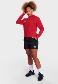 Rode zip-up hoodie met een kangoeroezak, gecombineerd met zwarte sportshorts met een logo. Model draagt zwarte sneakers en witte sokken.