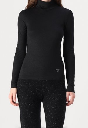 Kvinde iført en tætsiddende sort rullekrave og sorte bukser med prikker, stående med armene afslappet langs siden.