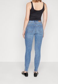 Pantalones vaqueros ajustados de mezclilla azul con una cintura alta, que presentan un diseño clásico de cinco bolsillos y un ligero desgaste. Top negro sin mangas y zapatos de charol.