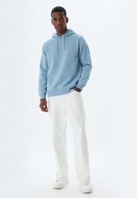 Sudadera azul claro con cordón y bolsillo tipo canguro, combinada con pantalones rectos blancos y zapatillas blancas. Textura de tela suave.