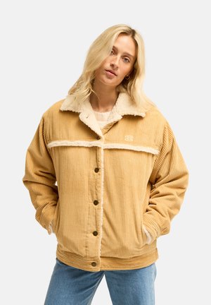Jeune femme en veste en velours côtelé beige avec une doublure en polaire blanche, les mains dans les poches, portant un jean bleu, debout devant un fond uni.