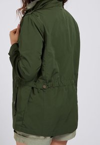 Oliwkowa zielona kurtka ze ściągaczem w talii, z wysokim kołnierzem i metalowymi guzikami. Wykonana z trwałej, gładkiej tkaniny.
