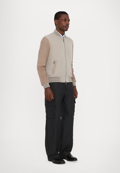 Hellbeige Bomberjacke mit kontrastierenden, tanfarbenen Ärmeln, Reißverschlusstaschen und gerippten Bündchen, kombiniert mit schwarzen Cargohosen.