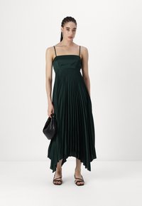 Mörkgrön plisserad klänning med fyrkantig halsringning, tunna axelremmar och asymmetrisk nederdel. Modellen håller en svart väska och bär svarta sandaler med remmar.