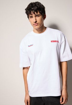 Vit bomull t-shirt med oversize-design, med röd och blå text "Potsdamer Platz" och en liten grön etikett vid fållen.
